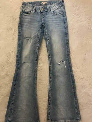 Ljusblå bootcut jeans med slitningar - Snygga ljusblå bootcut jeans med slitningar på benen och broderade bakfickor. Jeansen har klassisk femficksdesign, låg midja och stängs med knapp och dragkedja. Perfekta för en avslappnad och trendig look.