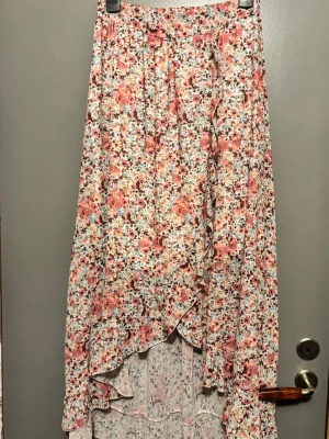 Blommig omlottkjol i pastellfärger från sportshopen - Säljer en lång omlottkjol med blommigt mönster i rosa, vit, blå och beige. Kjolen har volangkant och knytband i midjan för en skön och justerbar passform. Perfekt för dig som gillar romantisk och söt stil.