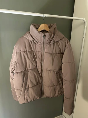 Beige pufferjacka med huva - Säljer en beige pufferjacka med dragkedja framtill och två sidofickor. Jackan har en stor huva med dragsko och quiltad design som ger en riktigt trendig look. Perfekt för kalla dagar och enkel att matcha med olika outfits.