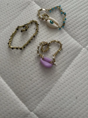 Halsband med snäcka och blå pärlor - Unikt halsband med en vit snäcka som hängsmycke och små blå pärlor längs den guldfärgade kedjan. Perfekt för dig som gillar strandvibbar och färgglada detaljer. Materialet är metall och snäcka, och pärlorna ger en extra touch.