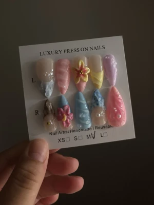 Lyxiga pastell press-on naglar - Handgjorda press-on naglar i pastellfärger med 3D-blommor och swirl-design. Färgerna är rosa, blå, gul, beige och lila. Naglarna har både glansig och matt finish samt olika former, bland annat stiletto och almond. Perfekt för dig som vill ha unika och kreativa naglar.
