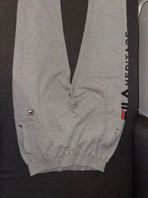Gråa mjukisbyxor från Fila  - Snygga gråa mjukisbyxor från Fila Heritage med logga och text längs benet. Byxorna har resår i midja och benslut samt fickor på sidorna. Perfekta för chill dagar eller träning. Tillverkade i mjukt bomullsmaterial för maximal komfort. Storlek Xl men sitter mer som L