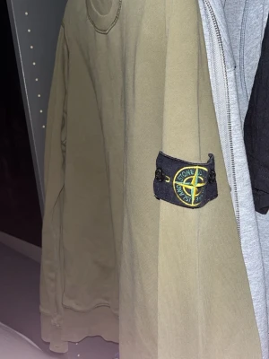 Olivgrön hoodie från Stone Island - Säljer en olivgrön hoodie från Stone Island med den klassiska loggan på vänster ärm. Hoodien har en avslappnad passform och är tillverkad i mjukt bomullsmaterial. Perfekt för dig som gillar streetwear och vill ha något stilrent med ikonisk badge. Storlek M-S