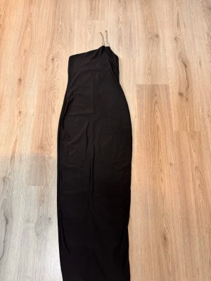 Svart oneshoulder långklänning - Stilren svart långklänning med oneshoulder-design och slits baktill. Klänningen har en smal passform och är tillverkad i ett mjukt, stretchigt material som ger en snygg siluett. Perfekt för dig som vill ha en enkel men elegant look.