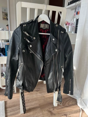 Svart skinnjacka från Jofama - Snygg svart skinnjacka från Jofama med flera dragkedjor, axelklaffar och bälte i midjan och är figursydd. Jackan har en cool bikerstil och rödsvart mönstrat innerfoder. Jackan är i fint skick men har blivit för liten för mig:)