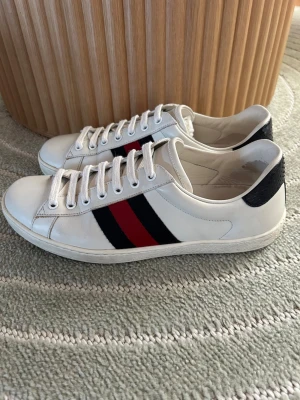 Gucci vita sneakers med röd/svart rand - Säljer ett par klassiska vita sneakers från Gucci med svarta och röda ränder på sidan samt svart detalj med Gucci-logga på hälen. Skorna har vita snören och är tillverkade i läder. Perfekta för dig som vill ha en stilren och lyxig look.