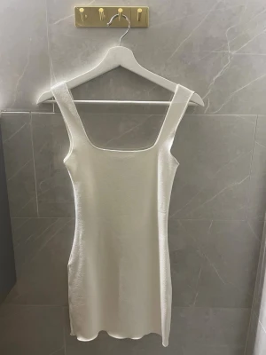 Vit kort tyg klänning från Zara - Säljer en stilren vit kort klänning från Zara med breda axelband och fyrkantig urringning. Klänningen har en figurnära passform och är tillverkad i ett mjukt material som ger en clean look. Perfekt för sommarens alla tillfällen!