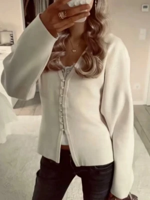 Krämvit kofta med knappar - Trendig krämvit kofta med v-ringning och dekorativa tygklädda knappar framtill. Koftan har lång ärm och en något figurnära passform. Perfekt att styla med jeans eller kjol för en snygg look.