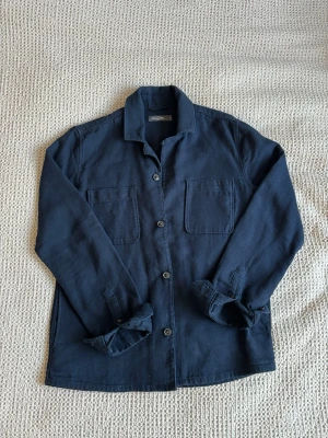 Mörkblå overshirt från Dressmann - Stilren mörkblå overshirt från Dressmann med två bröstfickor och knäppning framtill. Overshirten har klassisk krage och är tillverkad i ett mjukt, vävt material som ger en avslappnad look. Perfekt att bära öppen eller stängd över en t-shirt.
