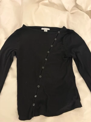 Svart asymmetrisk topp från Monki - Säljer en svart långärmad topp från Monki med asymmetrisk knappdetalj framtill. Tröjan har en smal passform och rund halsringning. Materialet är ribbat och stretchigt, vilket ger en skön och följsam känsla.