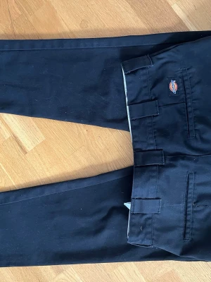 Svarta chinos från Dickies - Säljer ett par svarta chinos från Dickies med klassisk rak passform och diskret logga ovanför bakfickan. Byxorna har bälteshällor, två sidofickor och två bakfickor. Materialet är slitstarkt och passar perfekt till en clean och stilren look.