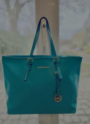 Turkos väska från Michael Kors - Snygg turkos toteväska från Michael Kors med guldfärgade detaljer och logotyp framtill. Väskan har dubbla axelband och en rund MK-berlock som hänger på framsidan. Tillverkad i slätt konstläder med stilren och rymlig design.
