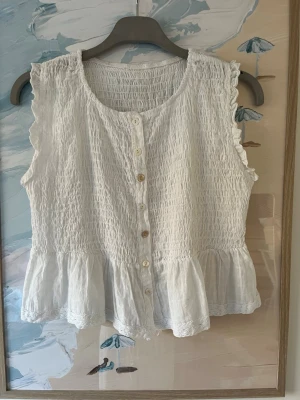 Vit ärmlös blus med volangkant - Söt vit blus med knappar framtill och volangkant nedtill. Blusen är ärmlös och har en smockad struktur som ger en skön och stretchig passform. Perfekt till sommaren och lätt att matcha med jeans eller kjol.