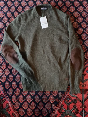 Olivgrön stickad tröja Barbour - Säljer en olivgrön stickad tröja från Barbour med rund hals och ribbade muddar. Tröjan har snygga bruna mockadetaljer på armbågarna och är tillverkad i mjukt material. Perfekt för dig som gillar klassisk och stilren look.