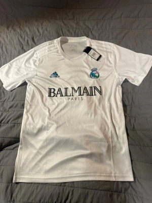 Vit Adidas x Balmain fotbollströja - Säljer en vit fotbollströja från Adidas i samarbete med Balmain Paris. Tröjan har Real Madrid-logga på bröstet, Adidas-logga i blått och Balmain-tryck på magen. Tillverkad i lätt och ventilerande material, perfekt för träning eller match.