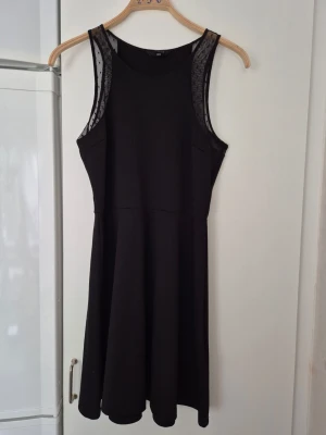 Klänning - Snygg svart klänning med meshdetaljer vid axlarna och ärmlös design. Klänningen har en enkel, figurnära överdel och utställd kjol. Perfekt för dig som gillar stilrena plagg med en twist.