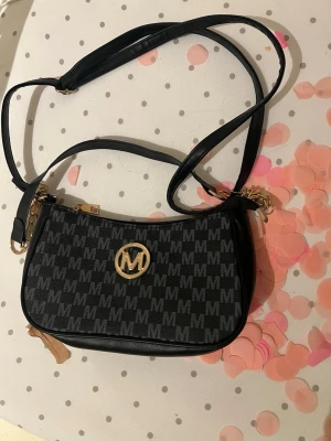 Svart väska   - Cool svart handväska med M-mönster över hela väskan och guldfärgad logga framtill. Väskan har en lång axelrem och detaljer i guld, perfekt för att lyfta din outfit. Materialet är syntetiskt och väskan har en stilren, rektangulär form.