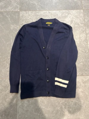 Mörkblå cardigan från Ralph Lauren  - Stilren mörkblå cardigan från Ralph Lauren med knäppning framtill och två vita ränder på ena ärmen. Väldigt eftertraktad tröja. Priset kan diskuteras vid snabb affär 