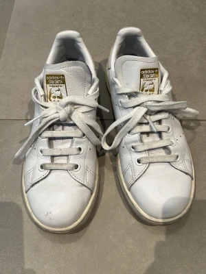 Adidas Stan Smith vita sneakers - Säljer ett par klassiska vita Adidas Stan Smith sneakers med beige häl. Skorna har rund tå, platta snören och är tillverkade i läder med gummisula. Perfekt för dig som gillar tidlös och clean stil.