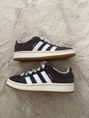 Adidas Campus svarta sneakers - Säljer ett par klassiska Adidas Campus sneakers i svart mocka med vita läderdetaljer och de ikoniska tre ränderna på sidan. Skorna är vit/begisa mellansula och gummisula i brunt. Mellan höga med snörning och diskret 'CAMPUS'-text på sidan. Har används max 2 gånger så de är som nya 