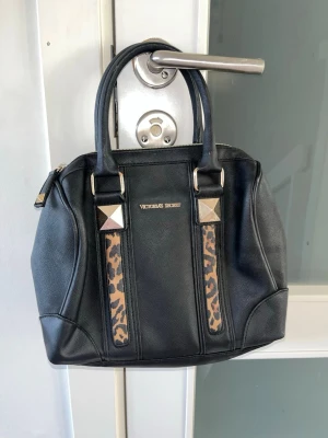 Svart handväska från Victoria's Secret - Snygg svart handväska från Victoria's Secret med leopardmönstrade detaljer och guldfärgade metalldekorationer. Väskan har två  handtag och dragkedja upptill🎀🎀