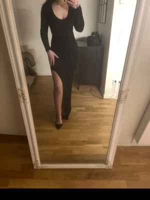 Svart långklänning med slits - Säljer en svart långklänning med djup v-ringning och lång ärm. Klänningen har en hög slits på ena sidan som ger en snygg och trendig look. Perfekt för dig som vill sticka ut! Materialet känns stretchigt och följsamt.