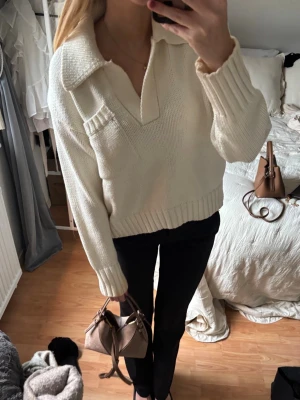 Vit stickad tröja med krage - Snygg vit stickad tröja med bred krage och djup v-ringning. Tröjan har en bröstficka och ribbstickade muddar vid ärmslut och nederkant. Perfekt för lager-på-lager och enkel att matcha med olika outfits.