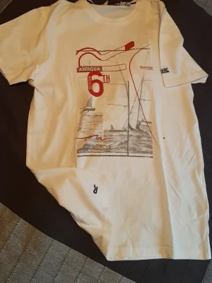Vit t-shirt Regatta med tryck - Vit t-shirt från Regatta Great Outdoors med coolt seglingsinspirerat tryck i rött och grått framtill. Motiv med texten 'Antigua 6th Edition' och segelbåtar. Klassisk passform och rund hals. Perfekt för dig som gillar marint tema. UNISEX. Storlek man: S/M, kvinna: M. Mått på bilderna.
