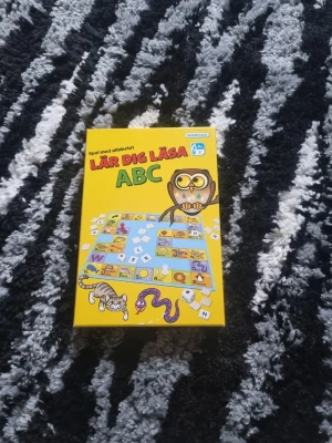 Lär dig läsa ABC spel - Säljer ett pedagogiskt brädspel från Kärnan, Lär dig läsa ABC. Spelet har en färgglad gul kartong med illustrationer av uggla, katt och orm. Innehåller bokstavsbrickor och spelplan som hjälper dig att lära dig alfabetet på ett roligt sätt.