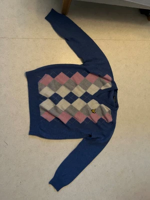 Blå argyletröja från Lyle & Scott - Snygg blå stickad tröja från Lyle & Scott med klassiskt argylemönster i rosa, vitt och grått på framsidan. Tröjan har V-ringning och den ikoniska gula fågeln broderad på bröstet. Perfekt för en preppy look och passar till jeans eller kjol. (En liten reva på andra bilden)
