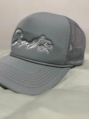 Grå Coreiz keps  - Snygg grå truckerkeps med silvrig brodyr framtill och mesh på sidorna och bak. Kepsen har böjd skärm och ett flätat band ovanför skärmen för extra detalj. Perfekt för en streetwear-look.