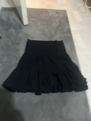 Svart volangkjol från 157 - Supersnygg svart kortkjol från 157 med bred smock i midjan och volangdetaljer. Kjolen har en flowy passform och är perfekt för dig som gillar en enkel men trendig stil. Passar till allt och är lätt att matcha med olika toppar.