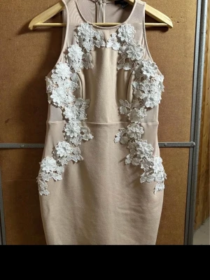 Beige klänning med spetsdetaljer - Snygg beige klänning med ärmlös design och vita blommiga spetsdetaljer framtill. Klänningen har en kroppsnära passform och dragkedja i ryggen. Perfekt för dig som vill sticka ut med en unik och elegant look.