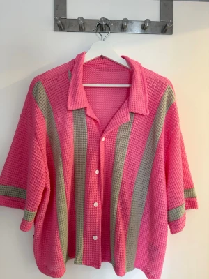 Rosa stickad skjorta  - Säljer en oversized stickad skjorta från Mamninty i rosa med breda gråa ränder. Skjortan har korta ärmar, krage och knäppning framtill med vita knappar. Materialet är grovstickat och ger en cool, avslappnad vibe.