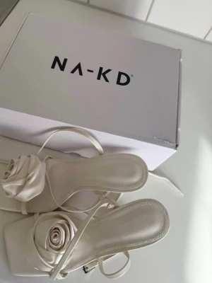 Vita klackar med rosettdetalj - Säljer ett par vita klackar från NA-KD med fyrkantig tå och eleganta remmar. Klackarna har en stor tygros framtill som ger en unik och trendig look. Tillverkade i satin material som känns lyxigt och passar perfekt till sommarens outfits.
