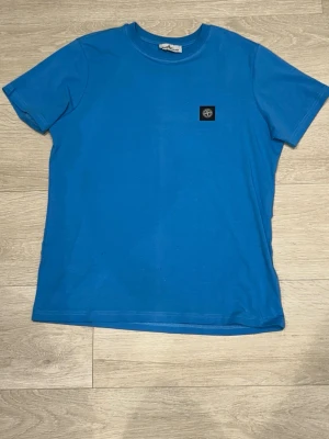Blå t-shirt från Stone Island - Snygg blå t-shirt  med klassisk rund hals och korta ärmar. T-shirten har en liten svart patch med logga på bröstet och är gjord i mjuk bomull för skön känsla. Perfekt till jeans eller shorts. Vet inte om det är äkta eller ej så därför de billiga priset! 