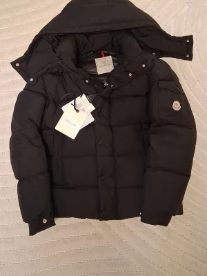 Svart dunjacka från Moncler - Snygg svart dunjacka från Moncler med avtagbar huva och klassisk Moncler-logga på ärmen. Jackan har stora fickor, dragkedja och tryckknappar framtill samt en ficka med lock på ärmen. Perfekt för kalla vinterdagar och riktigt trendig look. Använd ett fåtal gånger och har inga riktiga defekter😁