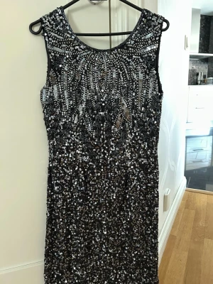 Glittrig svart klänning med paljetter - En riktigt snygg svart klänning med massor av glittrande paljetter i silver och guld. Ärmlös modell med rund halsringning och figurnära passform. Perfekt för dig som vill sticka ut och glänsa på festen. Materialet är troligtvis polyester.