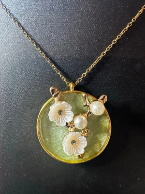 Guldigt halsband med blommor och pärlor - Unikt halsband med tunn guldfärgad kedja och runt hänge i grönt. Hänget har dekorativa vita blommor och två pärlor på en gren, allt inramat i guld. Perfekt för dig som gillar detaljer och naturinspirerade smycken.