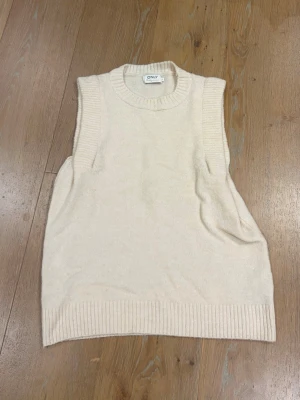 Beige stickad väst från ONLY - Snygg beige stickad väst från ONLY med ribbade kanter vid hals, ärmhål och nederkant. Perfekt att bära över en skjorta eller t-shirt för en trendig lager-på-lager-look. Enkel och stilren design som passar till mycket.