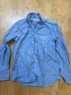 Blå skjorta från Brothers  - Snygg ljusblå skjorta från East West i klassisk modell med knappar framtill och krage. Skjortan är tillverkad i bomull och har långa ärmar. Perfekt för en stilren och avslappnad look. Köpt på brothers.