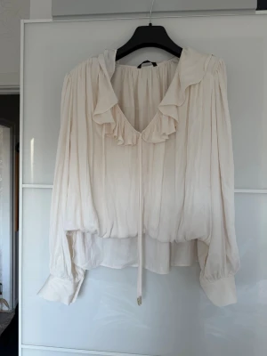 Krämvit blus med volangkrage - Säljer en krämvit blus med v-ringning och volangdetaljer runt kragen. Blusen har långa ärmar med breda muddar och ett tunt knytband framtill. Materialet är tunt och luftigt, vilket ger ett flowy intryck. Perfekt för en chic och avslappnad stil.