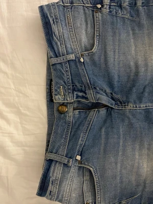 Blå jeansshorts från Lee - Snygga blå jeansshorts från Lee med klassisk femficksdesign, bälteshällor och knappgylf. Shortsen har en ljus tvätt och tydliga sömmar för en avslappnad look. Perfekta för varma dagar och enkel att matcha med olika stilar.