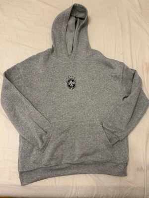 Grå hoodie med Saint-logga - Grå hoodie med broderad Saint-logga på bröstet, inspirerad av brasiliansk fotbollsstil. Klassisk känguruficka och huva. Mjuk och skön bomullsblandning, perfekt för en avslappnad look.