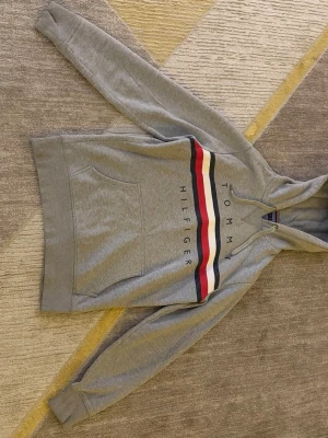 Grå hoodie Tommy Hilfiger M - Snygg grå hoodie från Tommy Hilfiger i storlek M. Klassisk design med huva och dragsko, stor magficka och breda ränder i rött, vitt och marinblått över bröstet. Tillverkad i mjuk bomullsblandning och har logotypen tryckt framtill.