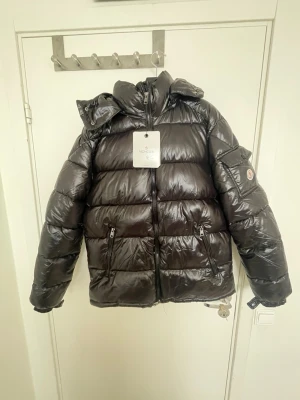 Svart pufferjacka från Moncler - Säljer en svart, glansig pufferjacka från Moncler med huva och dragkedja framtill. Jackan har två framfickor med dragkedja och Moncler-logga på ärmen. Perfekt för kalla vinterdagar och riktigt snygg streetstil.