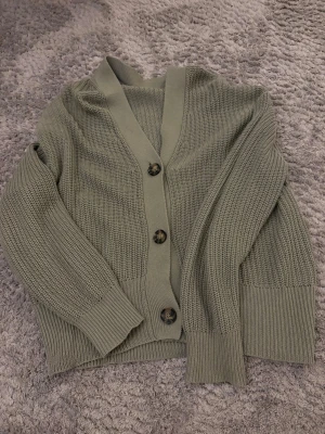 Olivgrön stickad kofta med knappar - Snygg olivgrön stickad kofta med stora, marmorerade knappar framtill. Koftan har v-ringning, ribbade muddar och bred kant nedtill. Perfekt för lager på lager och enkel att matcha med olika outfits. Har tvär klippt bort lappen så vet ej märket🥰