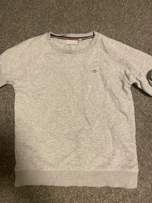 Ljusgrå sweatshirt från Gant - En stilren ljusgrå sweatshirt från Gant med rund halsringning och diskret Gant-logga broderad på bröstet. Tröjan har ribbade muddar vid ärmslut och nederkant samt klassisk passform. Tillverkad i mjukt bomullsmaterial för skön känsla. Passar folk mellan 154-160cm och den är väldigt fräsch. Nästintill helt ny tröjan.