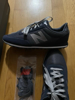 Svarta New Balance 410 sneakers 36,5 - Stilrena svarta New Balance 410 sneakers med grå detaljer och silverfärgade skosnören. Ovandelen är i mesh och syntetmaterial, med den klassiska N-loggan på sidan. Skorna har en låg profil och en vit sula med svart yttersula. Perfekta för en sportig och avslappnad look.