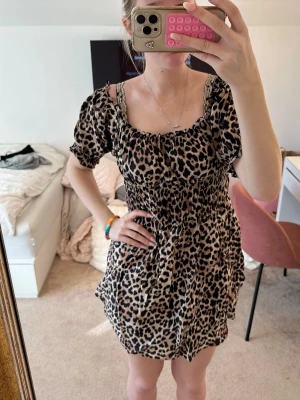 Leopardmönstrad kort klänning - Säljer en kort klänning med leopardmönster och puffärmar. Klänningen har smock i midjan och axelband samt en luftig kjol. Perfekt för dig som vill sticka ut med ett djurmönster och en avslappnad passform.